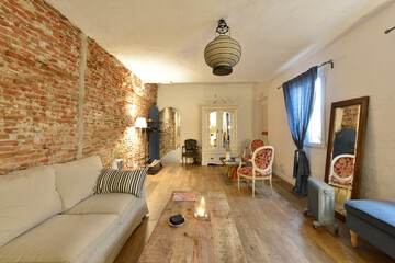 Living room with several chairs and armchairs, wooden floors, windows with blue curtains, exposed brick walls, mirror with a wooden frame leaning against the wall and wooden furniture