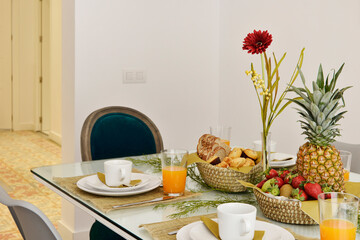 Dining table with glass top and wicker baskets with fresh fruits, sliced bread and sweet rolls with glasses of orange juice