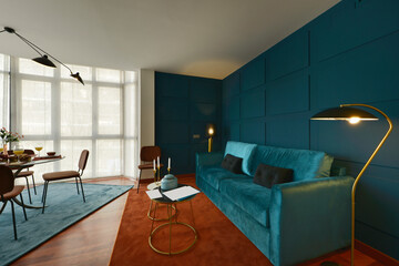 Living room in a studio apartment with a blue velvet three seater sofa, round glass and metal side tables, floor lamp and glass dining table and matching rugs