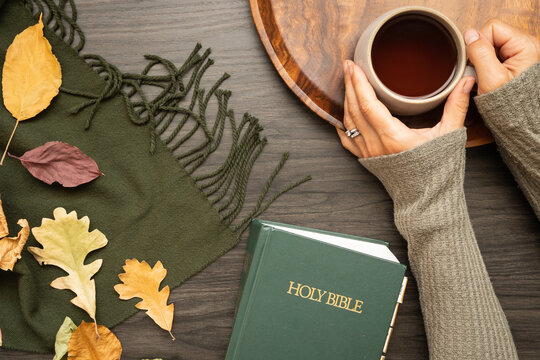 Hands of woman holding cup of coffee with bible and autumn leaves on a dark wood table