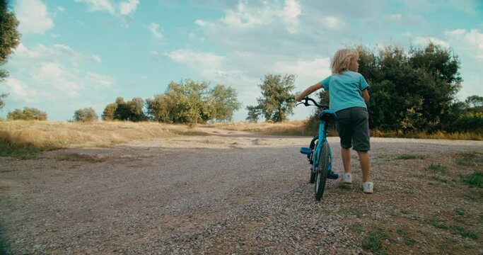 Happy carefree children walk on rural environment at summer vacation holding bicycle. Cheerful childhood concept. Following slow motion footage of child boy 