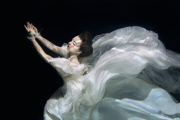 young woman in long white luxury dress underwater on the black background. Motion, art, winter...