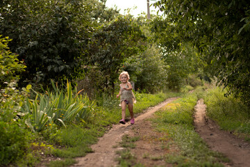 Fototapeta na wymiar A beautiful little Caucasian girl runs cheerfully and smiles along a country road in the summer in the village.