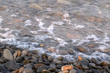 A clear transparent sea wave rolls over a rocky pebble beach, the concept of rest and travel, tranquility, relaxation and reflection on a warm summer day, close-up, selective focus, bokeh.