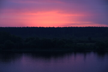 Fototapeta na wymiar A colorful sunset landscape with a purple sky above the black silhouette of a forest on the riverbank reflected in the lilac water