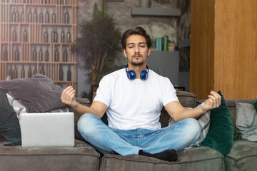 Young handsome man meditating during break. Freelancer doing yoga on couch after busy work day