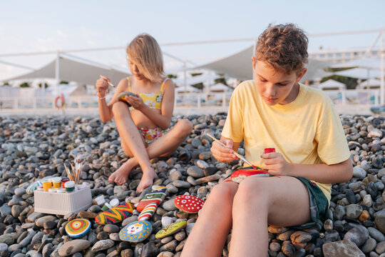 Children on the beach paint stones with paints and brushes.