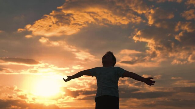 Little boy prays against background of sky and sun. Child raised his hands to sky in park at sunset, true faith. Religion and god, childhood dreams. Child plays in park against sky. Happy family