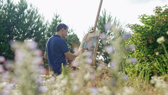 An artist paints a picture on an easel in a beautiful natural landscape. A man with a brush is painting surrounded by flowers. High quality 4k footage