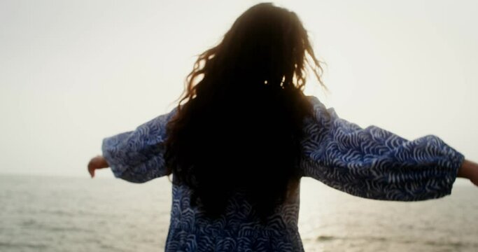 A woman smoothly moves her arms, spreading them to the sides, standing on the sea coast in the light of the setting sun and looking into the distance. Video filming from the back. 4k video, red komodo