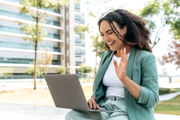 Online video conversation outdoors. Positive brazilian or hispanic young business woman, sitting outdoors with a laptop near the business center, talking with clients or coworkers by video call, smile