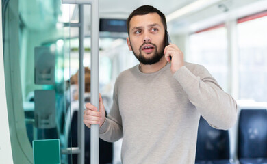Focused young bearded man talking on phone on his way to work in modern streetcar on spring day..