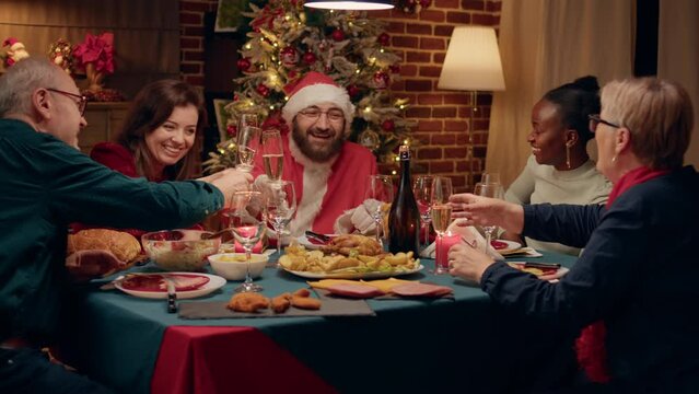 Joyful man disguised as Santa Claus clinking champagne glass with family enjoying Christmas dinner. Festive relatives celebrating winter feast while sitting at table with Santa Claus looking person.