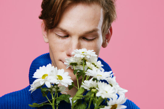  portrait of a handsome, guy with freckles on his face, in a blue sweater, sniffing a bouquet of large white daisies with his eyes closed in pleasure, standing against a pink background. studio photo