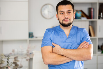 Portrait of confident male physician wearing surgical scrubs standing in his office with arms crossed and looking at camera.