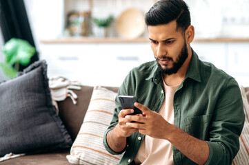 Puzzled indian or arabian guy in casual wear, sits on sofa in a living room, using smartphone,...