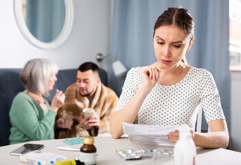 Portrait of dissatisfied young woman sitting at table choosing medicines for her sick husband, while her mother-in-law giving drugs to him