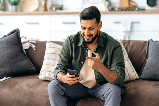 Happy positive arabian or indian guy in casual wear, spending time at home, sits on the sofa in the living room, using smartphone, chatting with friends, browsing the internet, social networks, smile