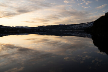 Magical view of the lake at sunset. The forest, dramatic sky and mountains in the horizon, reflecte din the water surface with a beautiful golden hour light.