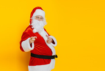 Fototapeta na wymiar A man in a santa costume points with his index fingers at the camera on a yellow isolated background with copy space.