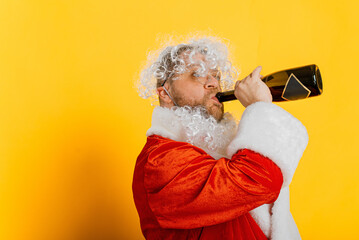 Drunk unshaven santa drinks champagne from the neck of a bottle on a yellow isolated background...