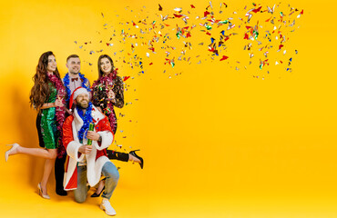 Confetti party. A group of friends in New Year's tinsel and a santa costume shoot a confetti popper on a yellow isolated background with copy space. Banner for new year party - obrazy, fototapety, plakaty
