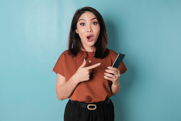 Surprised Asian woman wearing brown shirt holding her smartphone, isolated by blue background