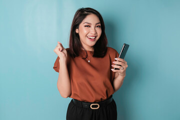 Fototapeta premium A young Asian woman with a happy successful expression wearing brown shirt and holding smartphone isolated by blue background