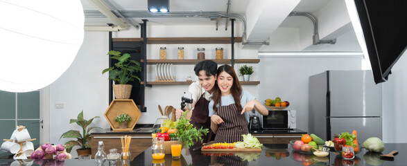 Asian couple spend time together in the kitchen. Young woman in apron cooking salad dish while his boyfriend recording vlog video for social blogger. Photography studio lights set up in the kitchen.