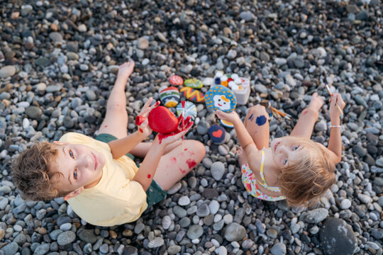 Brother and sister paint stones on the beach with paints and brushes.