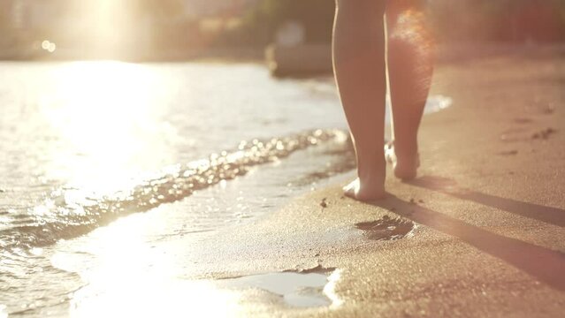 Close-up footage of female legs walking on the sandy beach at sunset near the sea. Ocean waves, sunlight, relaxation and recreation. High quality 4k footage