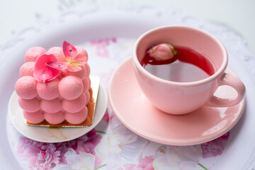 Serving festive tray of appetizing creamy cake with butterfly decoration and pink cup of fragrant tasty tea with flower petals top view closeup. Pink style decor, holiday, cute service