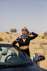 Fototapeta na wymiar A stylish girl in a black suit and a silk scarf enjoys a ride in a convertible car in the desert, Dubai, UAE
