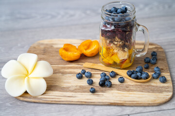healthy organic breakfast, transparent glass cup presentation, with mix of cut fruits and nuts: blueberries, berries, walnuts, apricot, peach, cranberry  on a wooden plate, frangipani flower decor