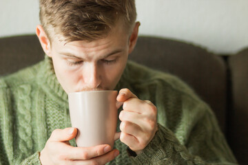 Caucasian man wearing green knitted woolen sweater drinking hot cup of coffee or tea at cozy home. Cold autumn morning.