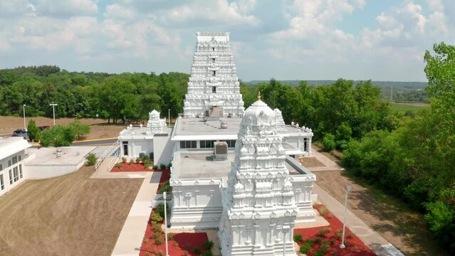 Aerial drone forward moving shot of a white hindu temple surrounded by green trees at daytime.