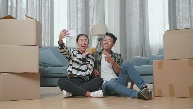 Young Asian Couple With Cardboard Boxes Sitting On The Floor And Taking Photo By Smartphone After Moving Into A New House 
