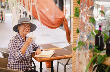 Attractive senior woman with hat sitting at cafe table reading a book while drinking an espresso coffee - caucasian lady enjoying free time and retirement