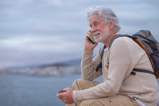 Smiling senior man sitting on the cliff in sea excursion smoking pipe looking at the horizon while talking on mobile phone. Caucasian grandfather with backpack enjoying vacation and freedom