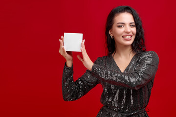 An elegant girl in a black short dress on a red background demonstrates an empty white box and looks at the camera. A beautiful brunette is holding a box with space for text. Discount advertising.