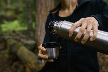 Woman rests alone in pine forest and drinking hot tea from steel thermos cup, natural moments and solo activity in nature