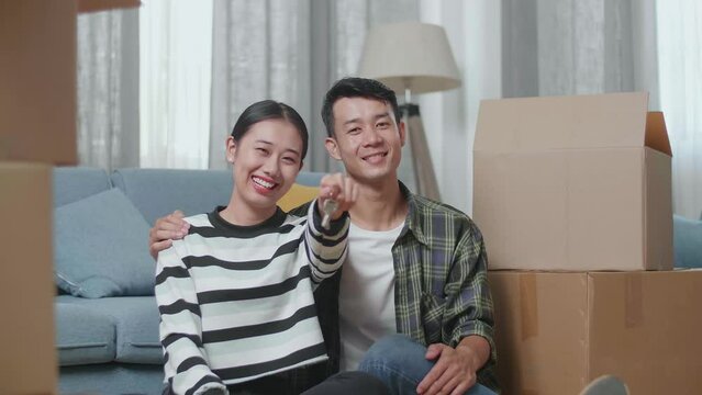 Young Asian Couple With Cardboard Boxes Sit On The Floor Smiling And Showing The Keys To Camera In The New House
