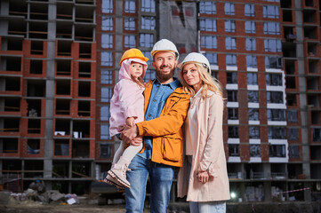 Fototapeta na wymiar Parents and daughter smiling while standing outdoors against apartment building under construction. Portrait of happy family homeowners at construction site.