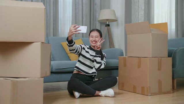 Young Asian Woman With Cardboard Boxes Sitting On The Floor And Taking Photo By Smartphone After Moving Into A New House 
