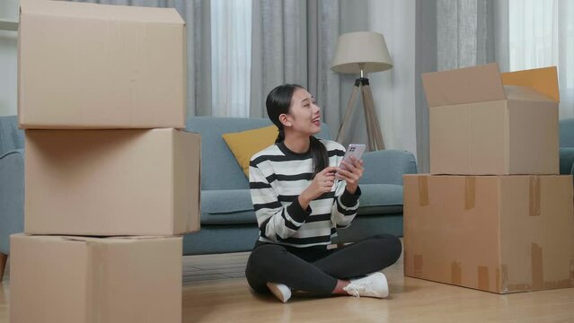 Young Asian Woman With Cardboard Boxes Sitting On The Floor And Comparing The House To The Photo On Smartphone After Moving Into A New House 
