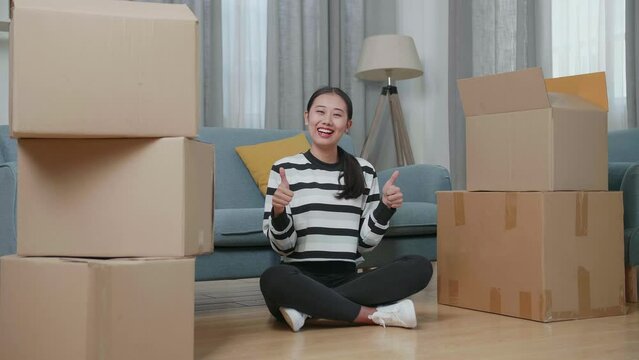 Young Asian Woman With Cardboard Boxes Sit On The Floor Smiling And Showing Thumbs Up Gesture To Camera In The New House

