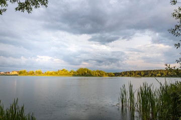 Sun during the rain. rainbow over the pond. Landscape with dramatic sky. Green plants near the water.