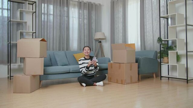 Young Asian Woman With Cardboard Boxes Sitting On The Floor And Comparing The House To The Photo On Smartphone After Moving Into A New House 
