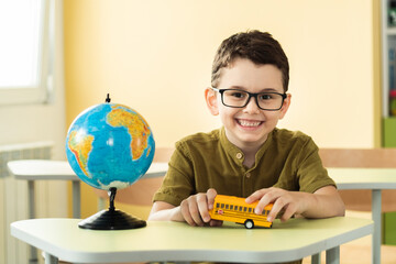 Cute caucasian schoolboy wearing glasses and sits at a desk in the classroom at elementary school. Little boy next the Globe playing with yellow school bus toy. Back to school concept