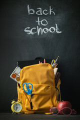 Naklejka premium Yellow backpack with school supplies next to the globe, red apple and glasses on the black school board background. Back to school concept on September day
